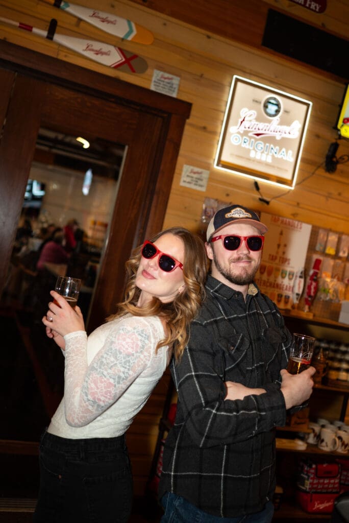 Brewery engagement photo shows couple wit fun sunglasses on and holding their drinks at the Leinie Lodge in Chippewa Falls, WI. Flash photo is true to color, by Maddie from Spirited Sage Photography, an Eau Claire photographer