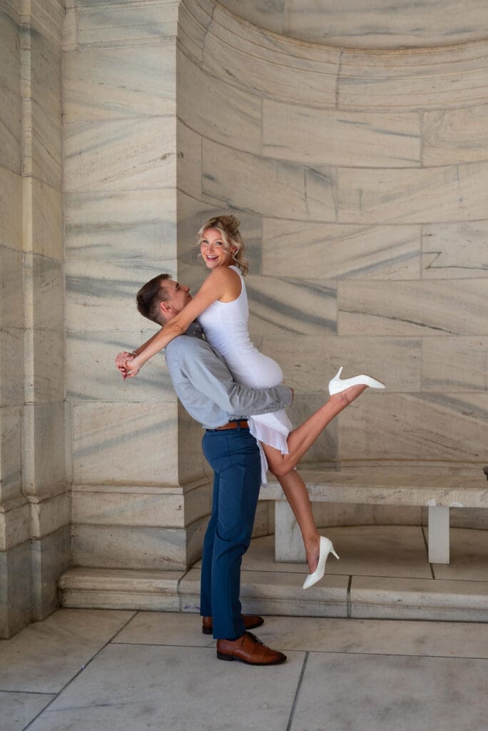 engagement photo at MN State capitol, showing man picking up his fiancé in her white dress and white shoes, photo by Maddie, a true to color Eau Claire wedding photographer