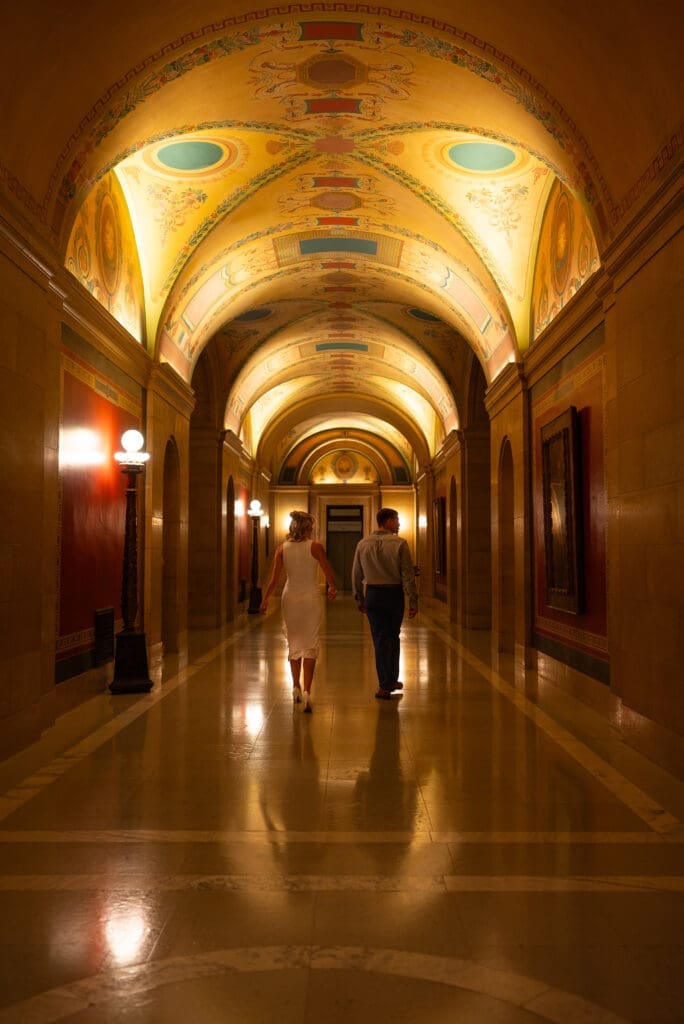 Artful and romantic shot of warm glow of lighting and art in MN State capitol and couple walking down the hall,. photographed in true color style by Maddie from Spirited Sage Photography