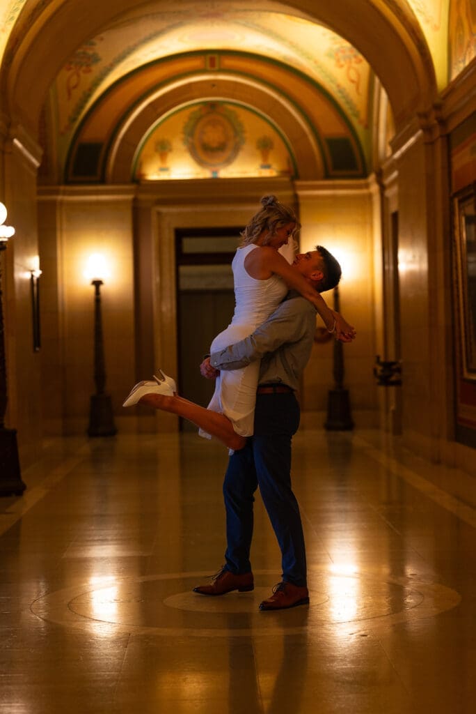 Man lifting and spinning his fiancé in the warm glow and art of the MN State Capitol for their St. Paul, MN engagement session. Maddie from Spirited Sage Photography is a true color Eau Claire Wedding photographer that offers a traditional or editorial