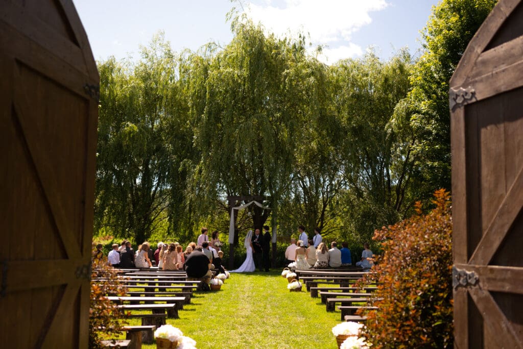 Ceremony at outdoor site with large doors, stained glass, and willow tree in Pepin, WI. Hidden Meadow and Barn wedding venue. Pepin photographer with availability.