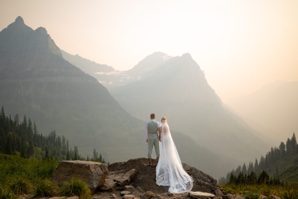 Bride and groom standing on large rock in Glacier National Park overlooking large mountain range and smoky skies