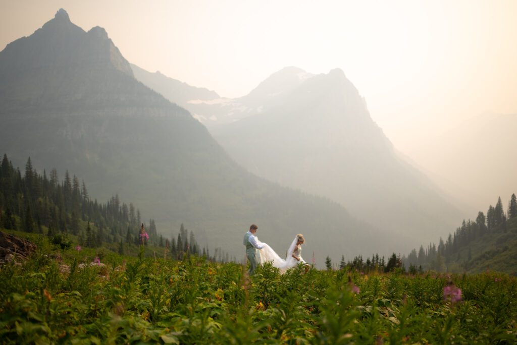 Stunning shot of bride and groom walking through the flowers of Glacier National Park with smoky and hazy mountains in the background.