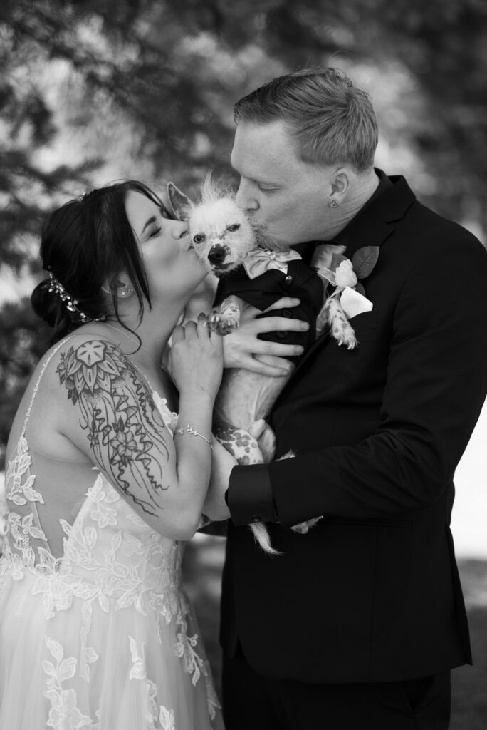 Tattooed bride and groom kissing hairless dog in tux on wedding day
